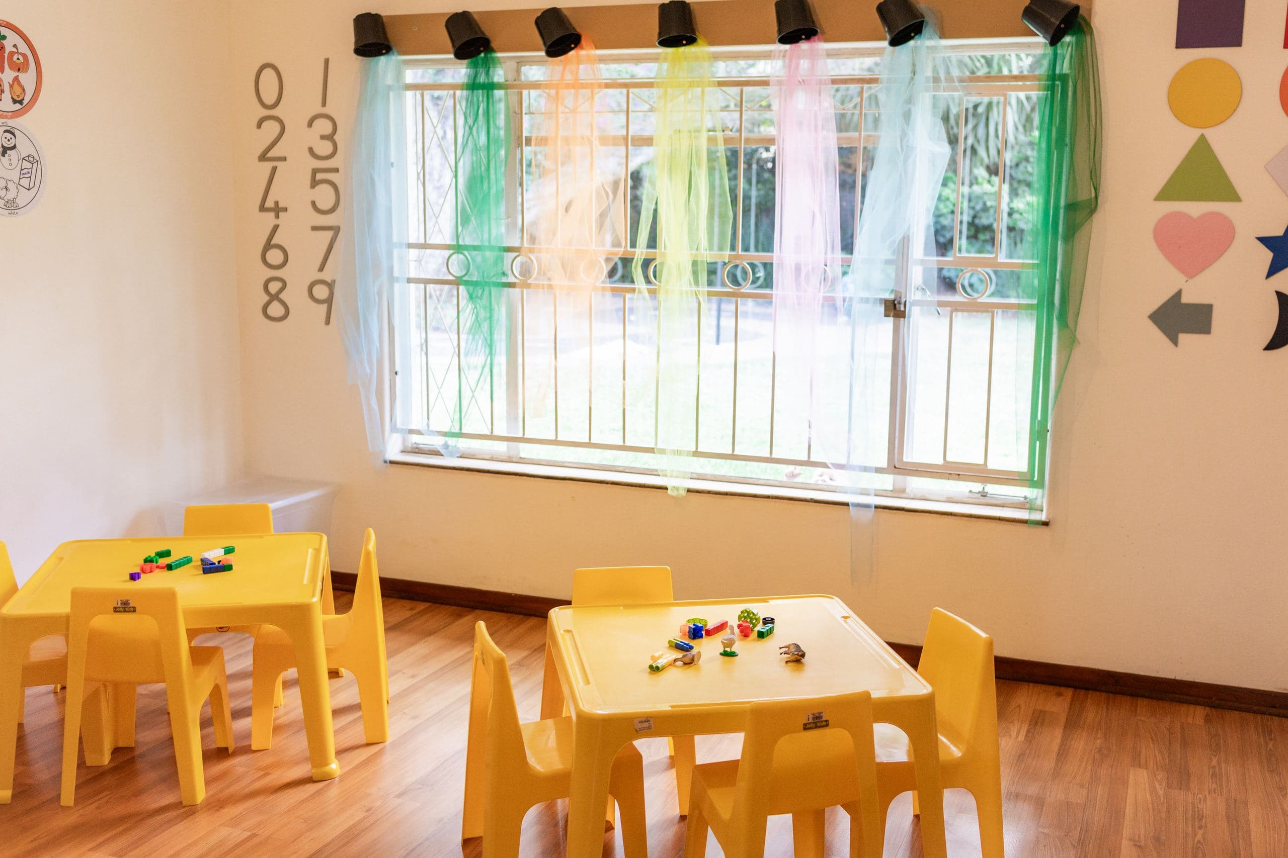 A classroom with yellow tables and chairs, a decorated window, and wall number and shape displays