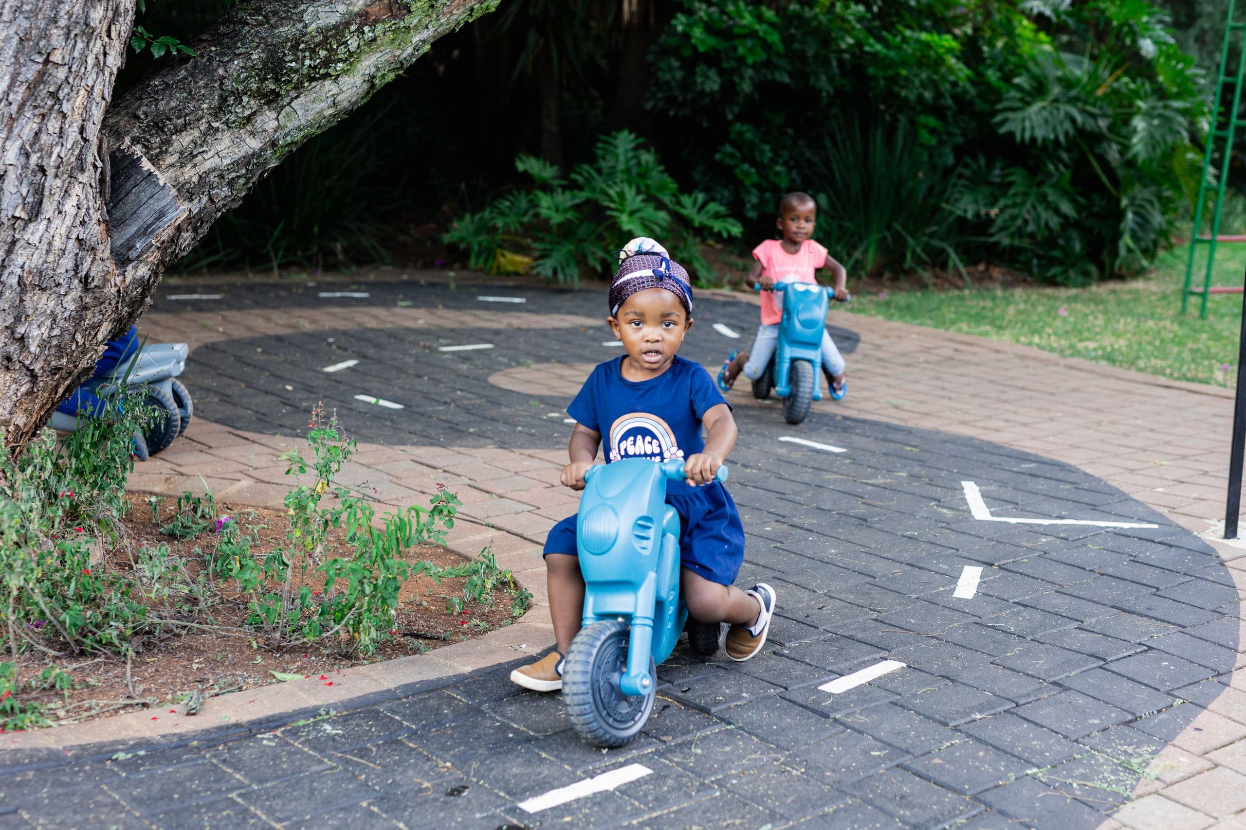 Two toddlers riding ride-on toy motorbikes along a paved outdoor path