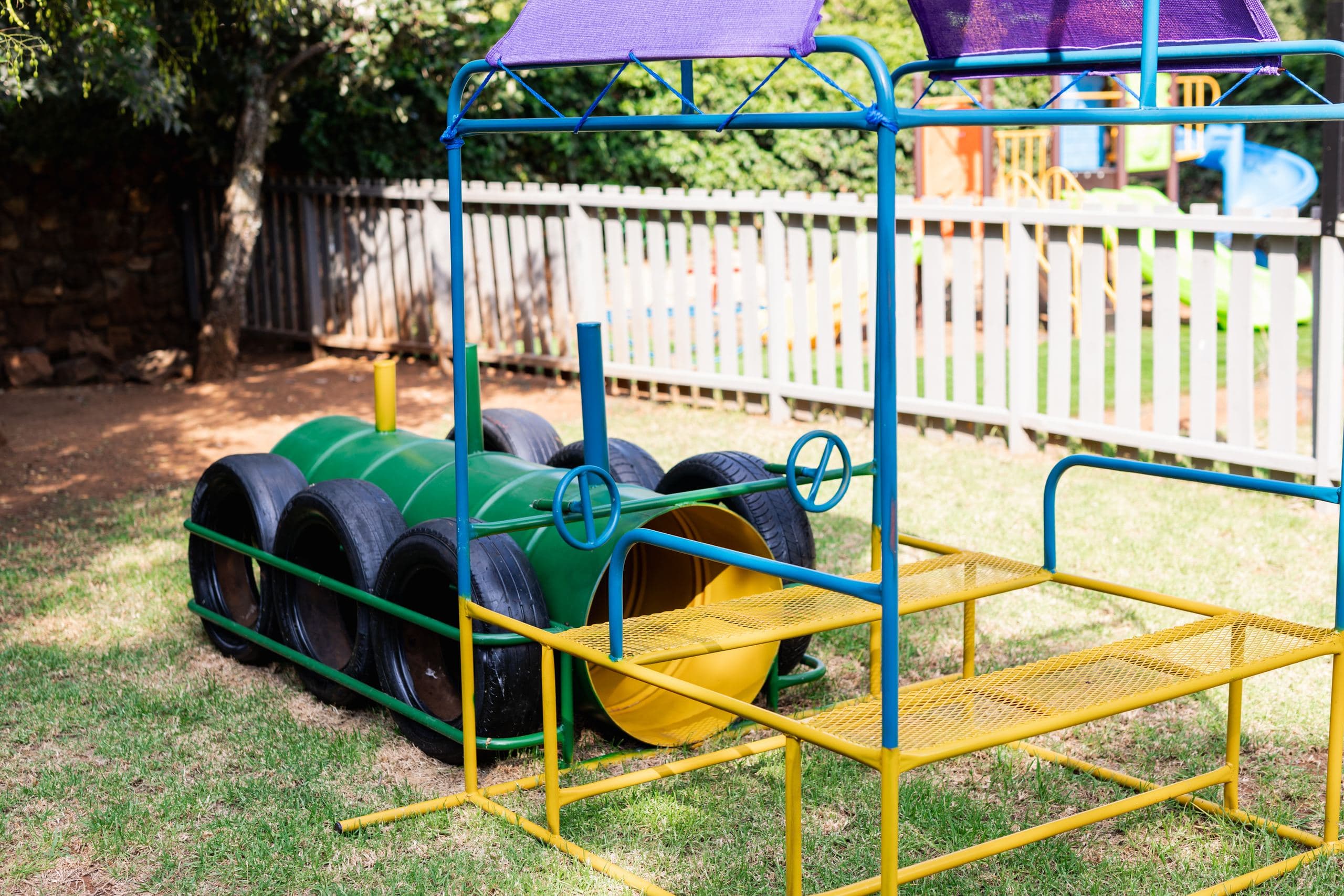Tyre-and-barrel obstacle play equipment on the lawn, with a wooden picket fence behind