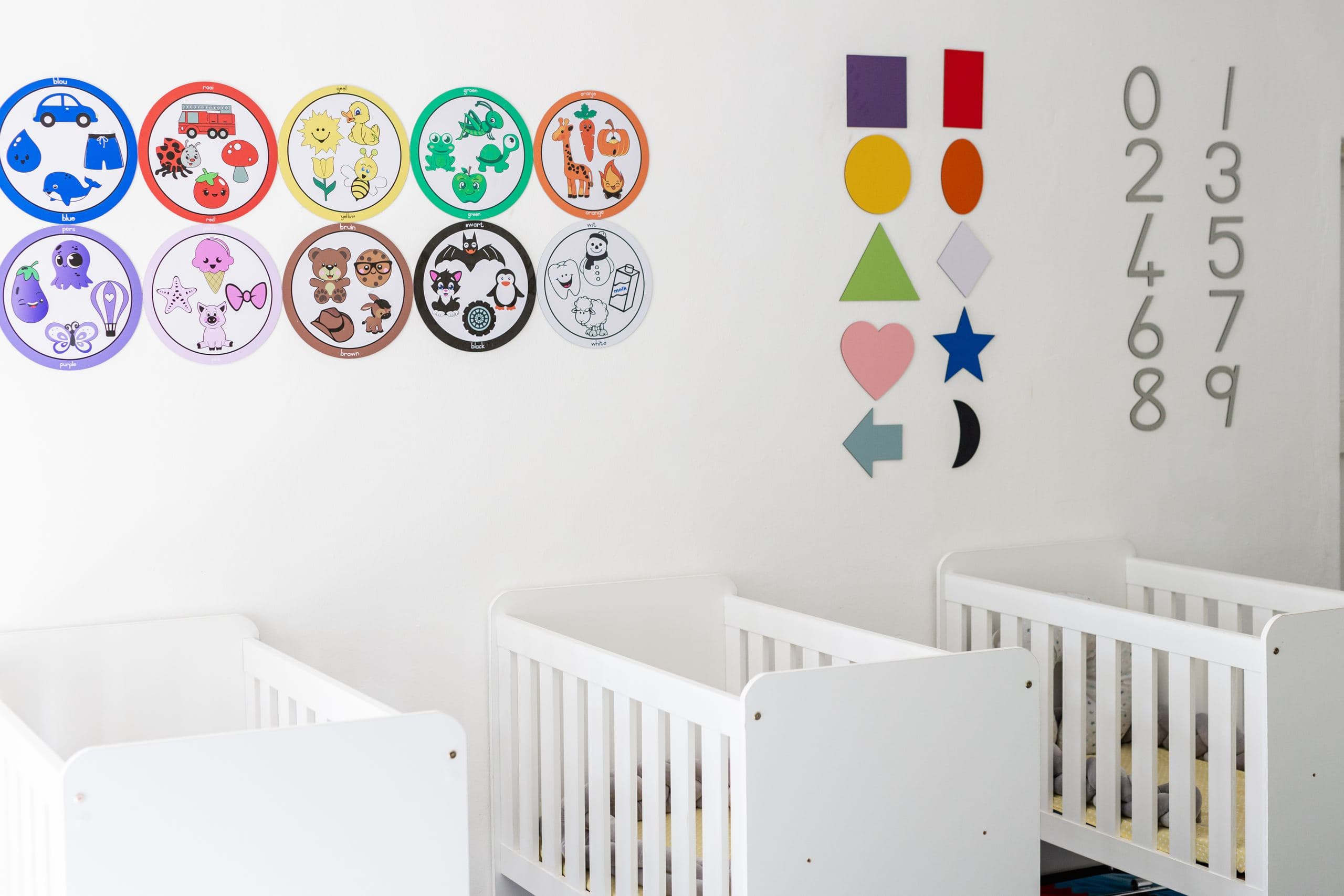 Bilingual colour-and-shape learning posters and wall numbers above cots in the baby room